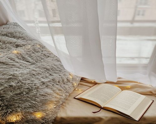 A cozy reading nook by a large window with soft sunlight filtering through sheer curtains in an Indonesian home
